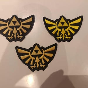 May include: Three embroidered patches featuring the Triforce symbol from The Legend of Zelda. Each patch is shaped like a winged crest with a black background and gold-colored thread forming the central triangle emblem and stylized wings.