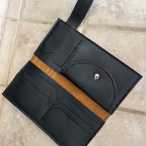 May include: A black leather wallet featuring a fold-over flap with a silver metal snap closure. The interior displays multiple card slots, a central pocket with a curved edge and a silver snap, and a visible tan suede lining with white stitching.
