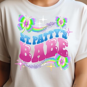 May include: White t-shirt with a colourful graphic. The design features the text "St. Patty's Babe" in blue and pink bubble letters, along with butterflies and rainbows. The butterflies have green, pink, and yellow wings.