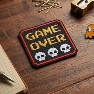 May include: A square embroidered patch with a black background, a red border, and yellow pixelated text reading GAME OVER above three white pixelated skulls. The patch is displayed alongside a metal pencil sharpener, pins, and a ruler.