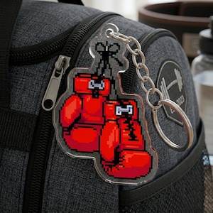 May include: A clear acrylic keychain featuring a pixelated design of two red boxing gloves. The keychain has a silver chain and a split ring. The background includes a gray duffel bag with a zipper and a weightlifting logo.