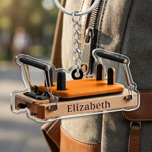 May include: An acrylic keychain shaped like a Pilates reformer machine with a light wood-toned base, an orange padded carriage, black shoulder rests, and metal hardware. The word Elizabeth is printed in black on the side of the wooden frame.
