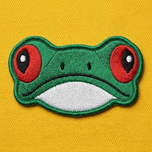 May include: An embroidered patch featuring the head of a red-eyed tree frog. The design displays a vibrant green face, large circular red eyes with black pupils and white highlights, two small black nostrils, and a white chin area with a black mouth line.