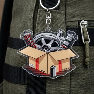 May include: A clear acrylic keychain featuring a cartoon illustration of car parts inside a cardboard box. The box contains a wheel, shock absorbers, and a red coil spring. A silver keyring is attached to the top. The text "UNBREAKABLE" is visible.