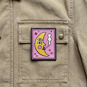 May include: A square patch featuring a yellow crescent moon with a face, smoking a cigarette, on a pink background. The patch is bordered in black and has small star and dot accents. The patch is sewn onto a tan jacket.