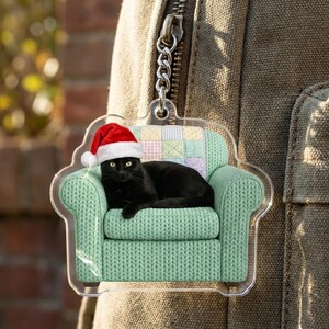May include: A clear acrylic keychain featuring a black cat wearing a red and white Santa hat, resting on a light green armchair with a textured knit pattern and a pastel-colored patchwork quilt backrest, attached to a silver metal chain.