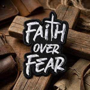 May include: A black embroidered patch featuring the white text "FAITH OVER FEAR" in a stylized, brush-stroke font. The word "FAITH" is positioned at the top with the letter "T" extending into a cross shape, followed by "OVER" and "FEAR" below.