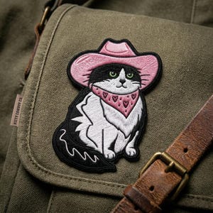 May include: Embroidered patch featuring a black and white cat wearing a pink cowboy hat and bandana with heart designs. The patch is on a green canvas bag, with a brown leather strap and a metal buckle visible.