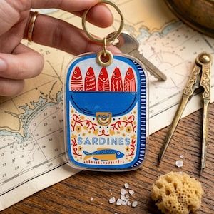 May include: A clear acrylic keychain featuring a blue sardine can design with the word "SARDINES". The can contains six red fish illustrations. The keychain has a gold-colored ring and is held by a hand.