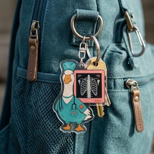 May include: A clear acrylic keychain featuring a cartoon goose in a teal medical uniform, stethoscope, and cap. The keychain also includes a pink x-ray image and keys, attached to a silver keyring. The keychain is hanging on a teal backpack.
