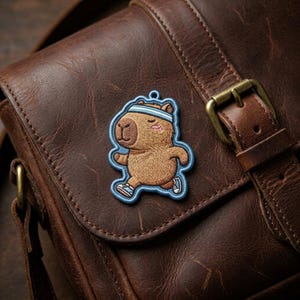 May include: Embroidered patch featuring a cartoon capybara running, wearing a blue headband and sneakers. The patch is brown with blue and pink accents, and is attached to a brown leather bag.