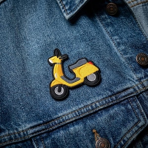 May include: An embroidered patch shaped like a yellow scooter with grey accents and black outlines, featuring a small red detail on the rear, attached to a blue denim jacket with visible metal buttons and stitched seams.