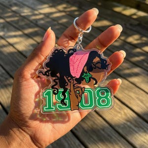 May include: A clear acrylic keychain featuring a stylized illustration of a woman with black hair and a pink head covering. The keychain has the number "1908" in green and white. A silver keyring is attached to the top.