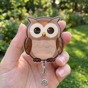 May include: A badge reel featuring a brown owl with large white eyes, dark brown wings, and a small yellow beak. The owl has a scalloped chest pattern and is attached to a clear plastic retractable cord mechanism with a metal clip at the bottom.