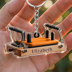 May include: A clear acrylic keychain shaped like a Pilates reformer machine. The miniature equipment features a light wood-toned base with the name Elizabeth printed on the side, an orange padded carriage, black shoulder rests, and metal hardware.