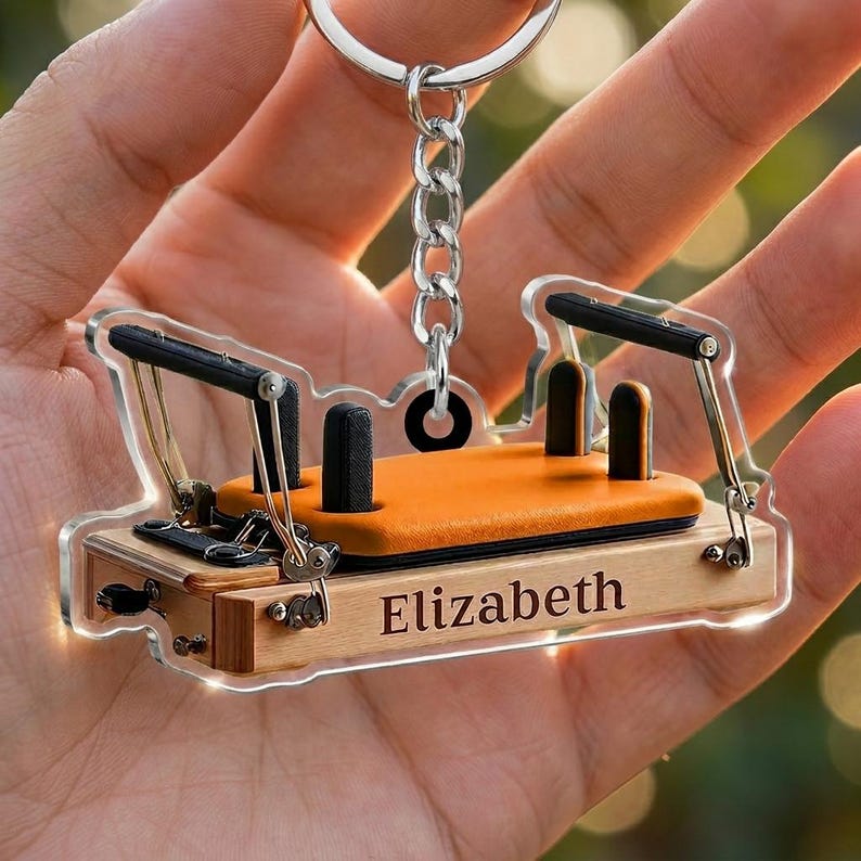 May include: A clear acrylic keychain shaped like a Pilates reformer machine. The miniature equipment features a light wood-toned base with the name Elizabeth printed on the side, an orange padded carriage, black shoulder rests, and metal hardware.