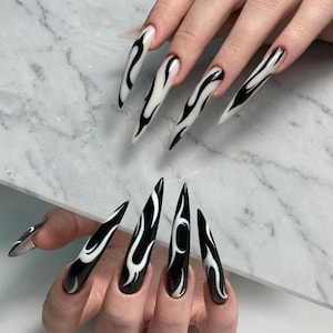May include: Long, pointed acrylic nails with a black base and white flame design. The nails are shown on two hands, with a marble background. The nail art is a bold and edgy style.