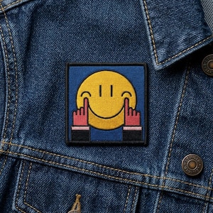 May include: A square patch featuring a yellow smiley face with two fingers pointing up on each side. The patch has a blue background and is sewn onto a blue denim jacket.