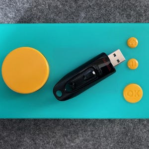 May include: A teal-colored rectangular tray with a large yellow circle and three smaller yellow circles. A black USB flash drive is placed on the tray. The smaller circles have icons: a house, a pause symbol, and the word "OK".