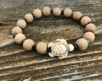 May include: A wooden bead bracelet with a white ceramic turtle charm. The beads are a light brown color and there are a few darker brown beads interspersed throughout the bracelet.