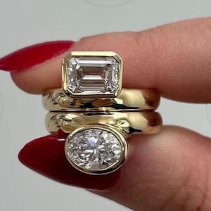 May include: Two gold rings stacked together, each featuring a bezel-set gemstone. The top ring holds a rectangular emerald-cut stone, while the bottom ring features an oval-cut stone. Both rings have smooth, polished yellow gold bands.