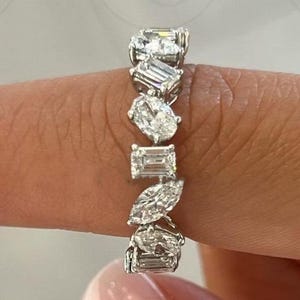 May include: A silver-toned band ring featuring a series of multi-shaped clear stones, including emerald, oval, and marquise cuts, arranged in a continuous pattern along the metal band, showcasing a variety of geometric facets and brilliant clarity.