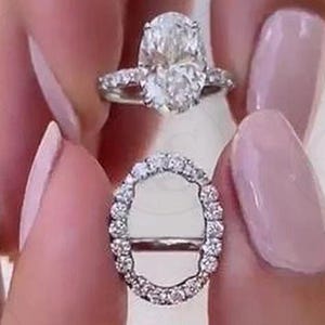 May include: Two diamond engagement rings. The top ring features a large oval-cut diamond with a halo setting and a diamond band. The second ring is a diamond band with an oval shape. Both rings are silver in color.