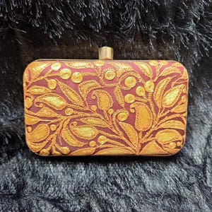 Könnte beinhalten: Eine rechteckige Clutch mit bordeauxfarbenem Grund und aufwendiger goldener Blumenstickerei. Die Clutch hat einen goldenen Verschluss und ist vor einem dunklen, strukturierten Hintergrund platziert. Das Design zeigt Blätter und paisleyartige Formen.