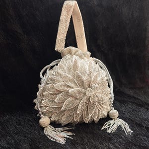May include: A silver beaded drawstring bag with a floral design. The bag has a beaded handle and tassels. The bag is made of a silver material and is decorated with intricate beadwork. The bag is on a dark background.