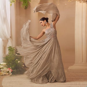 May include: A woman wearing a flowing, one-shoulder gown in a light taupe color. The dress features intricate beadwork on the bodice and a pleated skirt. She is holding a matching scarf, creating a dynamic pose. The background is a soft, neutral tone.