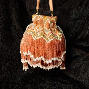 Könnte beinhalten: Eine pfirsichfarbene Perlenhandtasche mit Kordelzugverschluss und passendem Riemen. Die Tasche hat goldene und weiße Pailletten am oberen Rand und einen Besatz aus pfirsichfarbenen Perlen mit weißen Perlenakzenten.