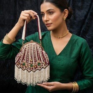 May include: An ornate, burgundy handbag with a pearl and bead design. The bag features a gold-colored frame, a burgundy velvet body, and a white pearl fringe. Green and gold embellishments add detail. The handle is white with burgundy trim.