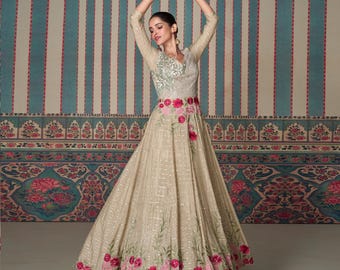 May include: A beige formal gown with a V-neckline and long sleeves. The bodice features intricate embroidery, and the waist is adorned with pink floral accents. The skirt flows gracefully, with matching floral detailing along the hemline.