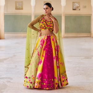 May include: A vibrant, multi-colored lehenga ensemble. The skirt features a tie-dye pattern in yellow, pink, and green. The cropped top is adorned with floral embellishments. A sheer, light green shawl completes the look. This traditional Indian outfit is perfect for festive occasions.