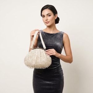 May include: A cream-coloured beaded handbag with a rounded shape and a clasp closure. The bag has a white strap and is held by a person wearing a dark grey velvet dress. The background is a plain white.