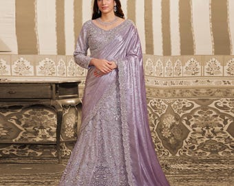 May include: A lavender saree with detailed embroidery and a matching long-sleeved blouse. The saree has a shimmering, draped fabric and is decorated with delicate embellishments. The outfit is suitable for formal events.