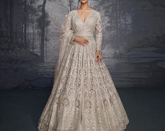 May include: A light beige, floor-length gown with intricate, embroidered detailing. The dress features a deep V-neck, long sleeves, and a fitted waist. A matching sheer shawl is draped over the arm. The gown is suitable for formal events.
