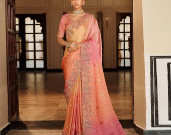 May include: A woman wearing a peach and pink saree with intricate embroidery. The saree features a gradient effect, transitioning from a light peach to a vibrant pink. The design includes detailed floral and geometric patterns.
