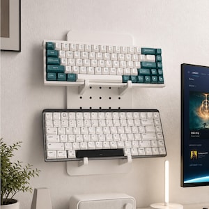 Puede incluir: Un estante de pared perforado de color blanco que sostiene dos teclados mecánicos. El teclado superior tiene teclas blancas y verde azulado oscuro, mientras que el teclado inferior tiene un marco blanco con teclas blancas y una barra espaciadora negra.