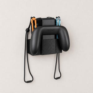 May include: A black plastic wall-mounted holder containing a black Nintendo controller grip. The grip features an orange wrist strap on the left and a light blue wrist strap on the right, both hanging down from the controller unit.
