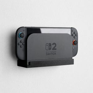 May include: A black Nintendo Switch console docked in a wall-mounted holder. The console features black Joy-Con controllers with one blue and one orange analogue stick. The dock displays the Nintendo Switch logo and text on its front surface.