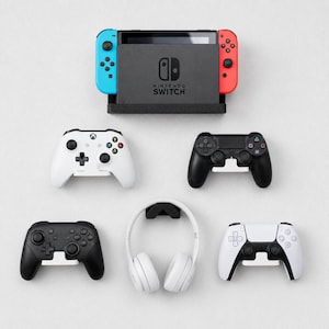 May include: A black Nintendo Switch console dock with a blue and red controller, accompanied by four various gaming controllers in black and white, and a pair of white over-ear headphones, all mounted on individual wall-mounted display stands.