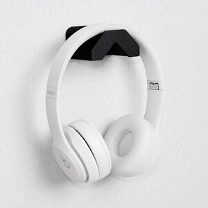 May include: A pair of white over-ear headphones resting on a black, V-shaped wall-mounted holder. The headphones feature a smooth white finish with a circular logo on the side, while the holder has a textured black surface and angular design.