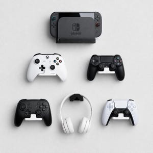 以下が含まれることがあります： 黒いドックに入ったNintendo Switch本体、白いXboxコントローラー、黒いPlayStationコントローラー、黒いProコントローラー、白いオーバーイヤーヘッドホン、白いPlayStation 5コントローラーが、それぞれ個別の壁掛けスタンドに設置されています。