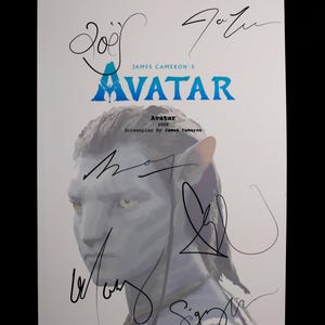 Op de afbeelding: Een wit papier met de titel "AVATAR" in blauw en de tekst "JAMES CAMERON'S" erboven. De afbeelding toont een blauwhuidig personage uit de film en meerdere handtekeningen met zwarte inkt.