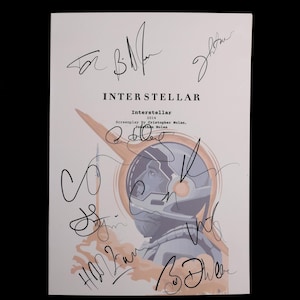 May include: A white poster featuring the title "INTERSTELLAR" with multiple signatures in black ink. The poster includes an illustration of an astronaut's helmet and the film's title and credits.