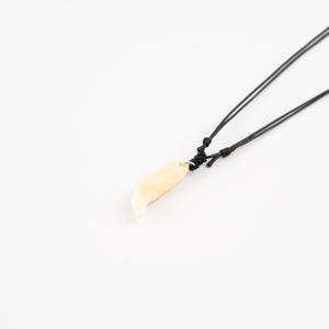 May include: A cream-coloured, tooth-shaped pendant necklace. The pendant hangs from a black cord, knotted at the top. The design is simple and striking. The background is plain white.