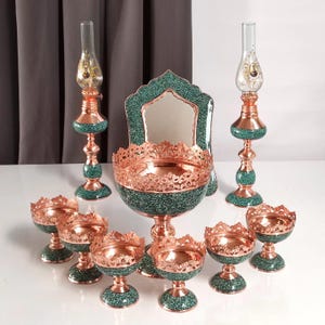 May include: A collection of decorative copper and turquoise items. The set includes two oil lamps, a mirror, a large bowl, and several smaller bowls. The copper pieces have intricate designs, and the turquoise elements add a pop of color. The items are arranged on a white surface.