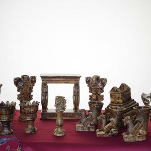 May include: A collection of decorative, brown, carved objects, including vases, pedestals, and architectural elements, arranged on a red surface. A single red rose in a glass vase is also present. The objects have a weathered, antique appearance.