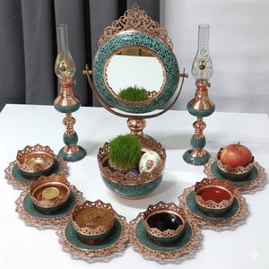 May include: A decorative table setting featuring a round mirror, two ornate lamps, and several small bowls and plates. The mirror and lamps have a copper-colored frame and teal accents. The bowls contain various items, and a red apple is also present.